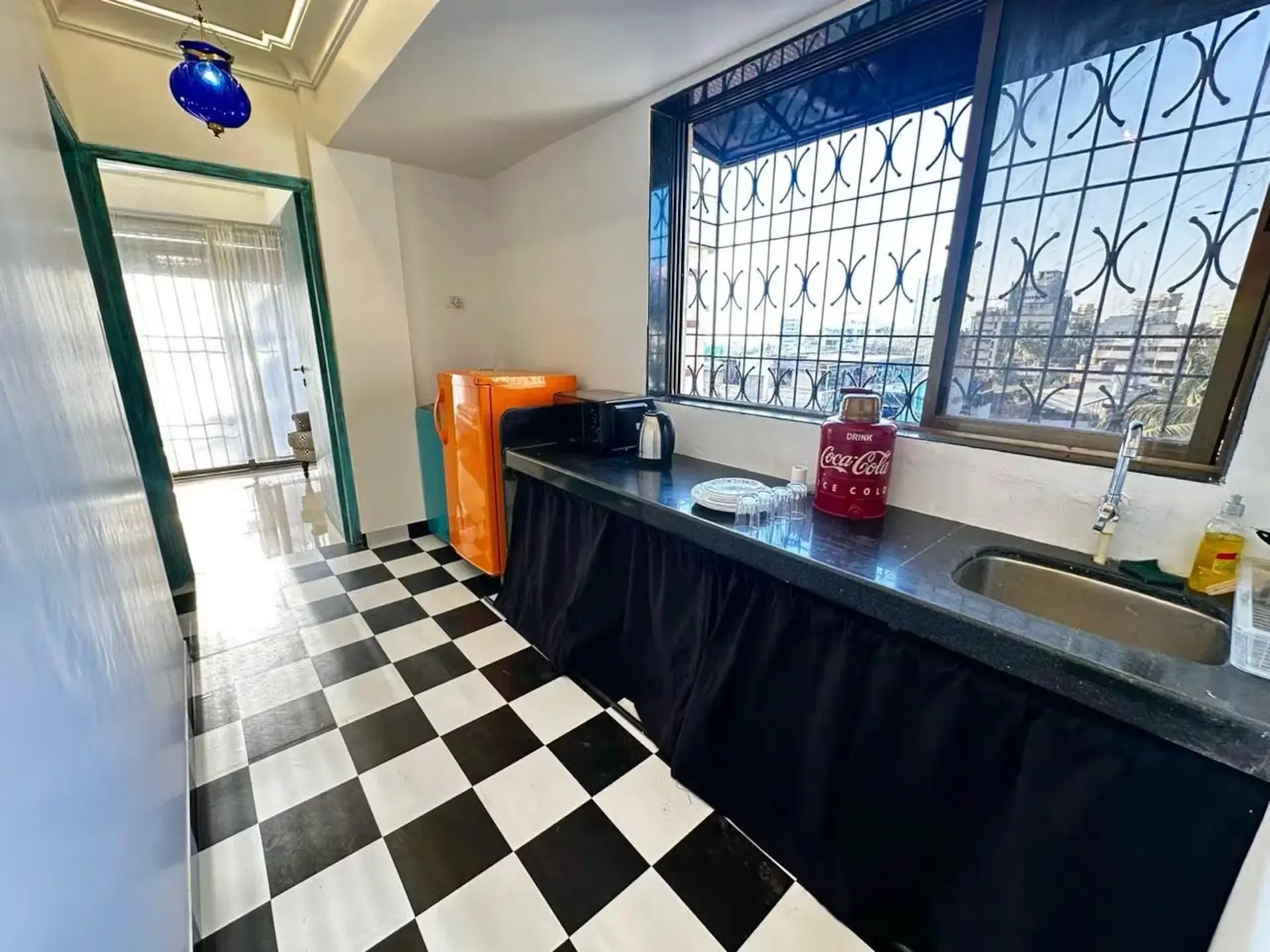 Kitchenette with a black-and-white chequered floor, an orange fridge and a window with an ornate grille