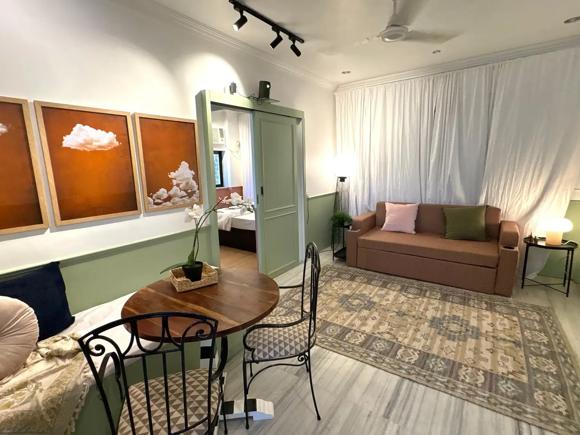 Living and dining area with a round wooden table, sage-green panelling and a sofa bed beneath cloud artwork