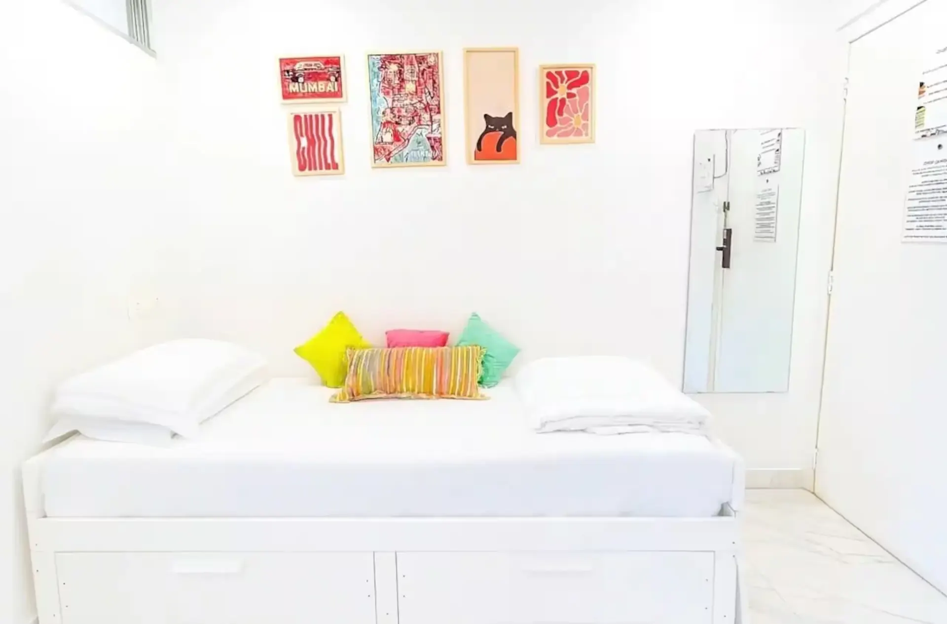 Bed with yellow, pink and green cushions beneath a row of colourful framed posters