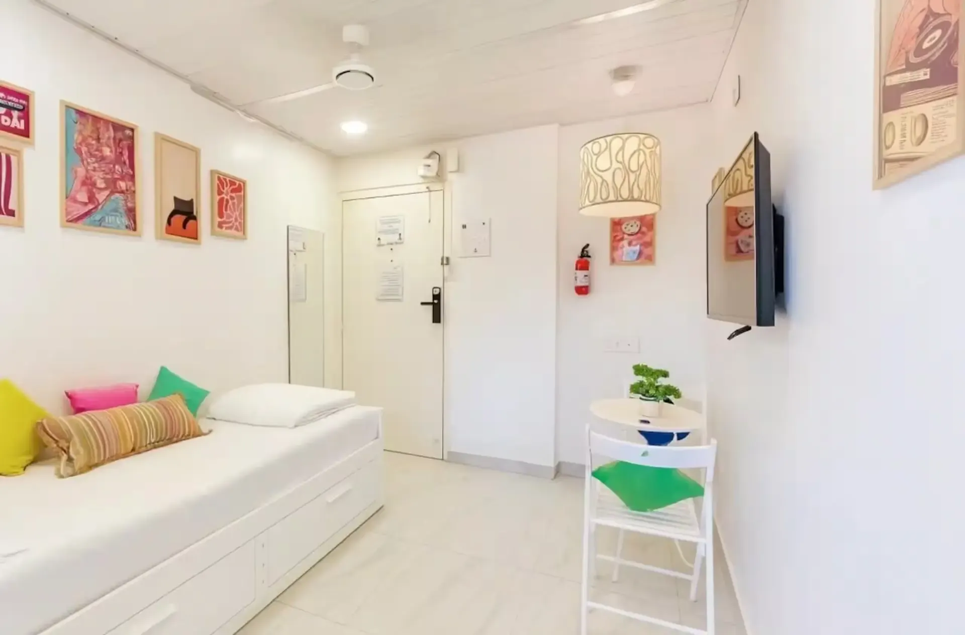 White studio seen from the bed, with posters, a patterned pendant lamp, a wall-mounted TV and the entrance door